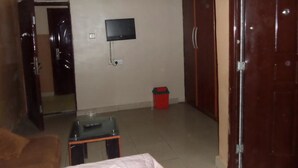 A room with a television mounted on the wall, a small table, a red trash bin, and a wardrobe. - B & S Hotel (Lagos)