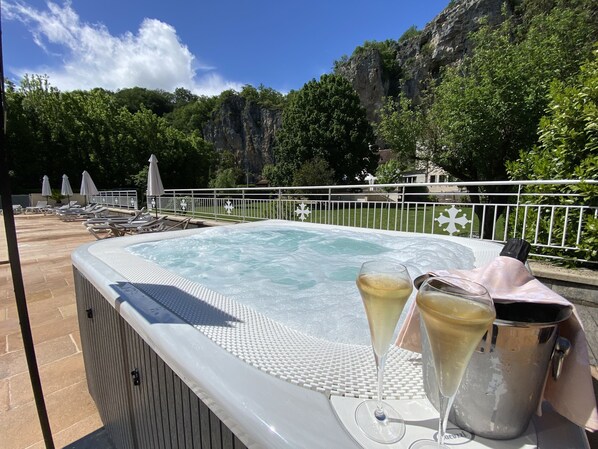 Un jacuzzi con sillones de descanso y sombrillas, rodeado por una valla blanca, con montañas al fondo.