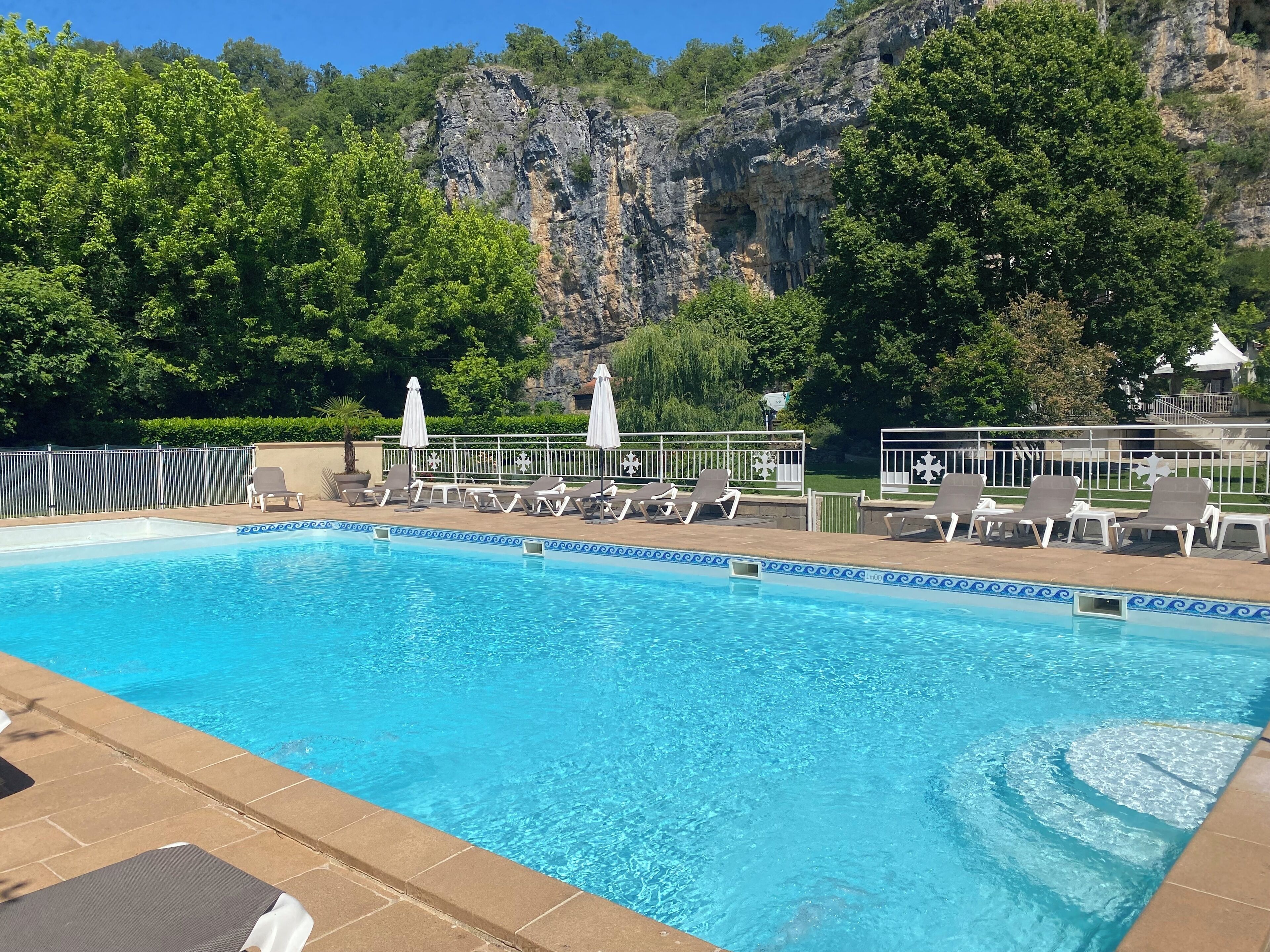 A swimming pool with lounge chairs and umbrellas, surrounded by greenery and a rocky cliff.