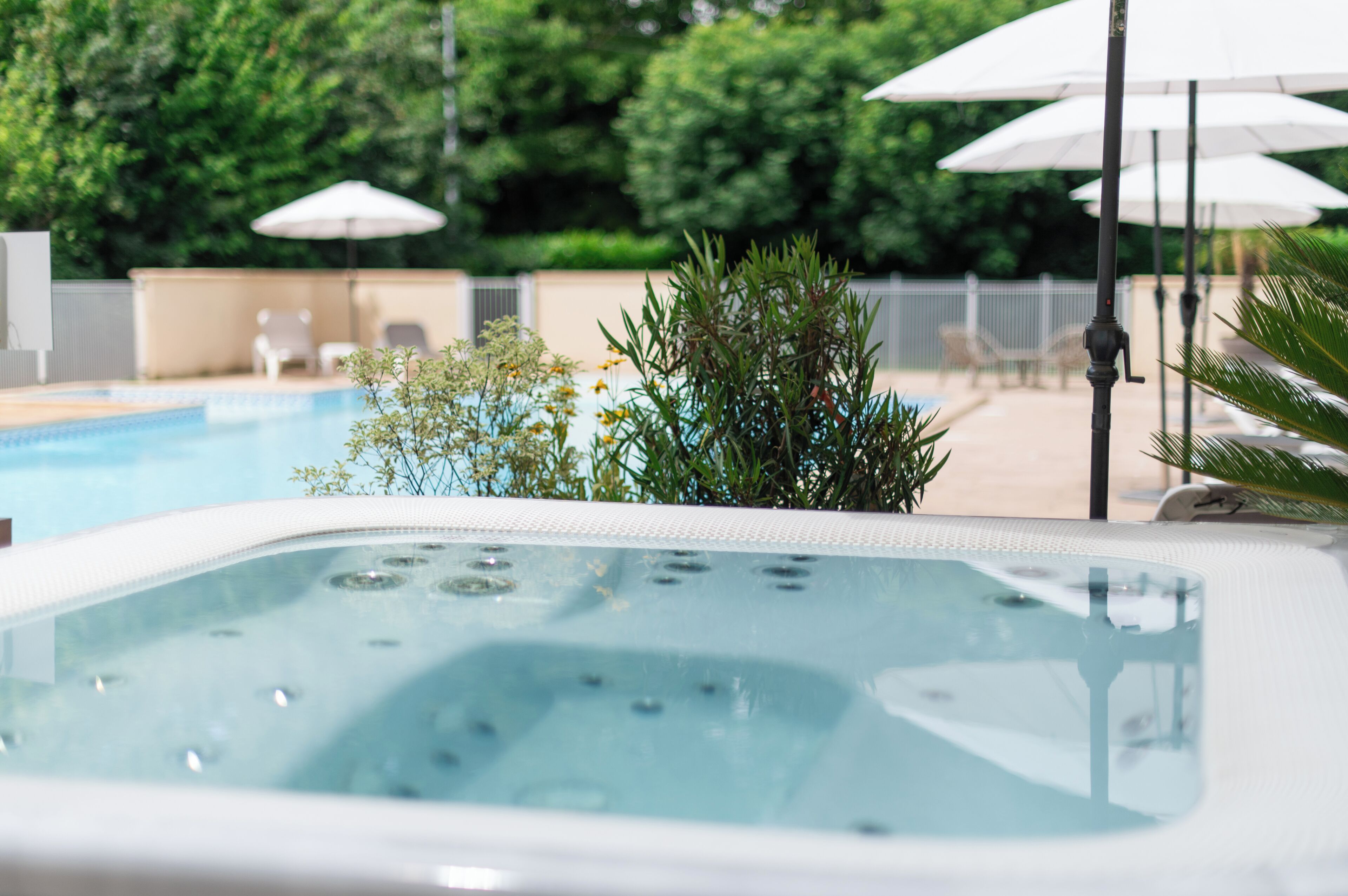 A hot tub with a pool in the background, surrounded by greenery and outdoor seating.