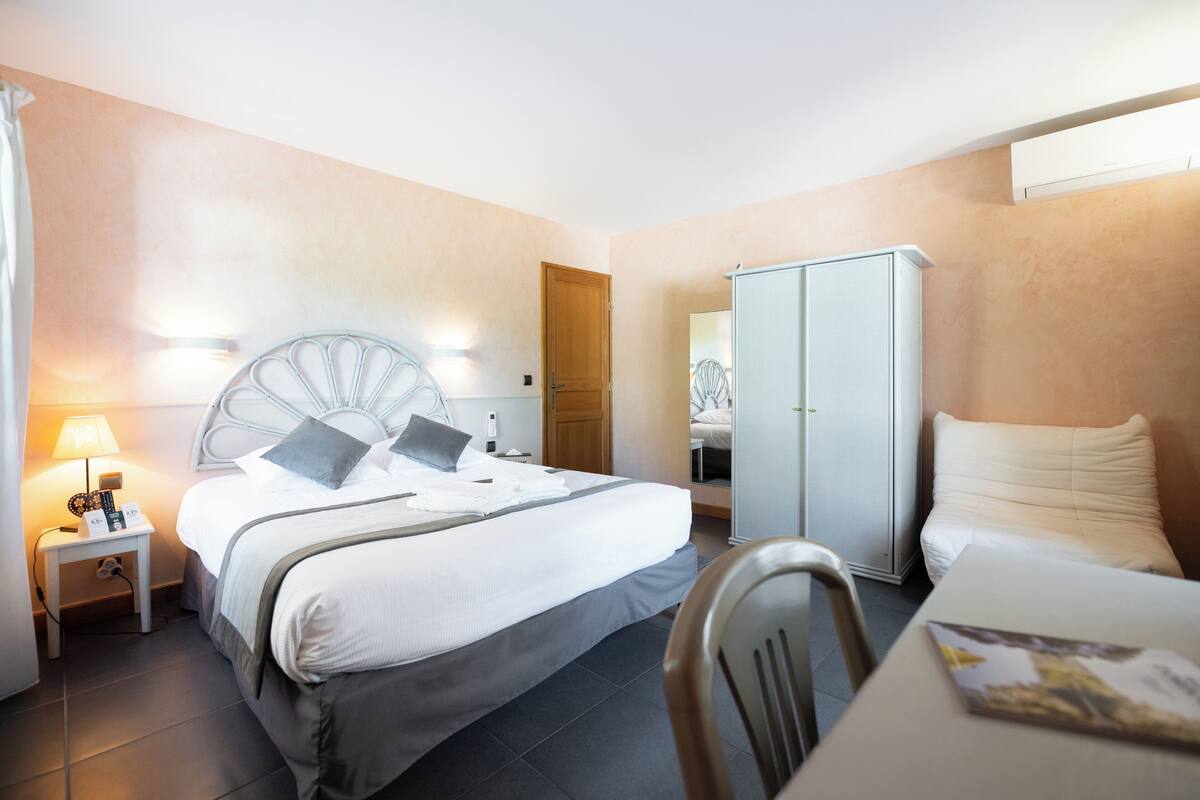 A hotel room with a bed, bedside table, lamp, wardrobe, and a chair.