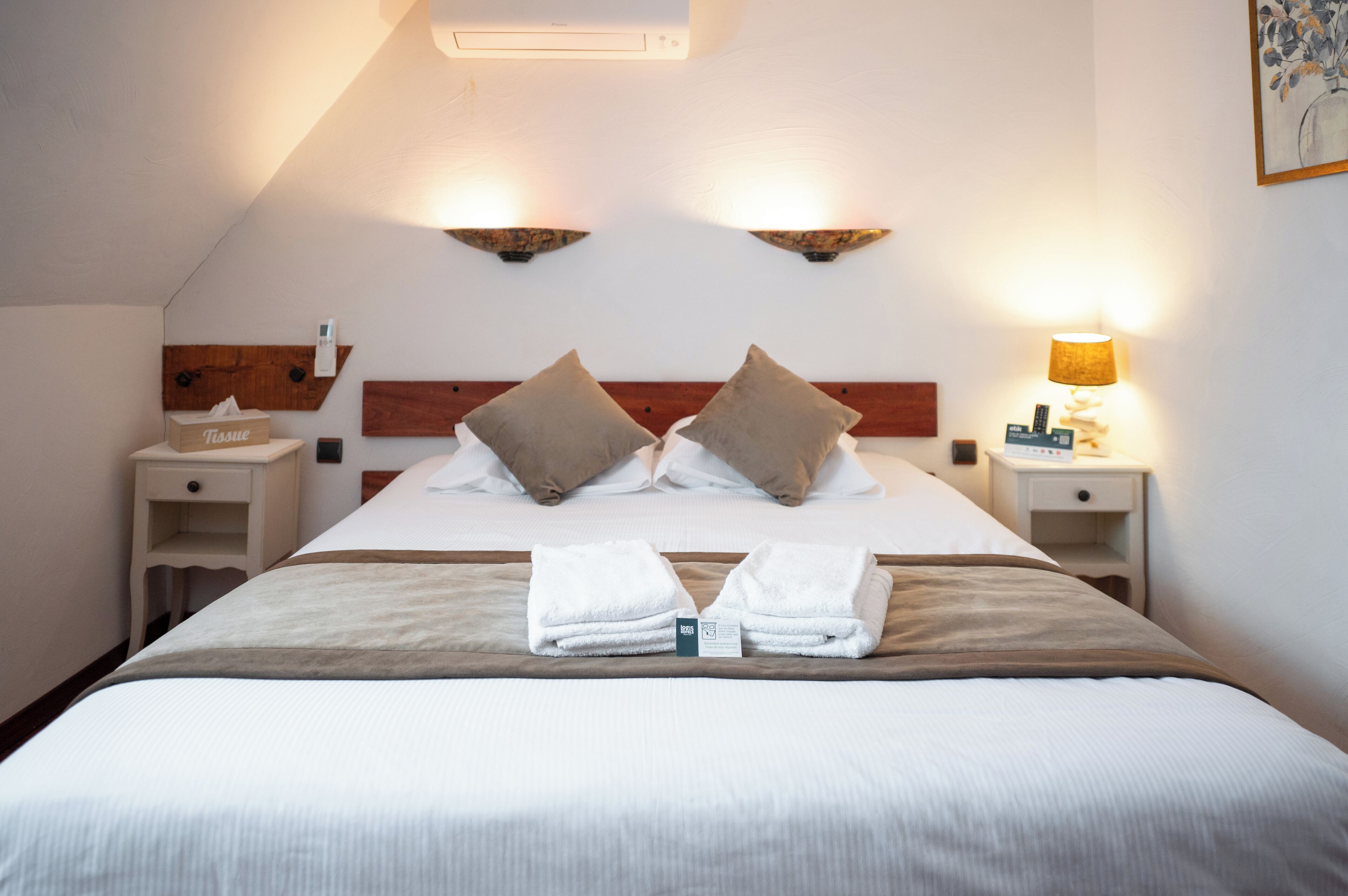 A neatly made bed with two pillows, a bedside table with a tissue box, and a lamp on the right.
