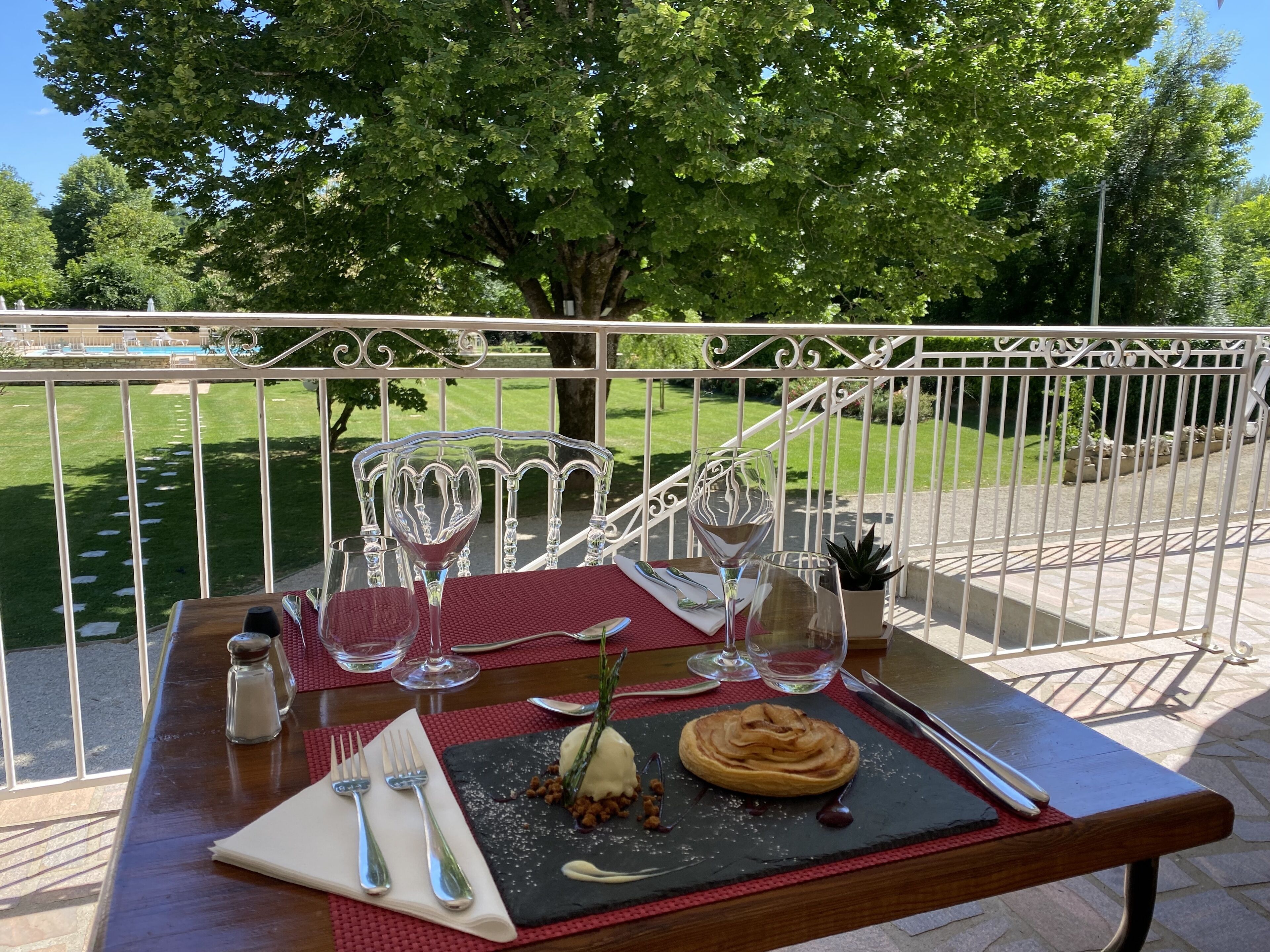 Mesa de comedor al aire libre con postre, copas de vino y cubiertos, con un jardín y una valla blanca de fondo.