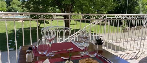Mesa de comedor al aire libre con postre, copas de vino y cubiertos, con un jardín y una valla blanca de fondo.