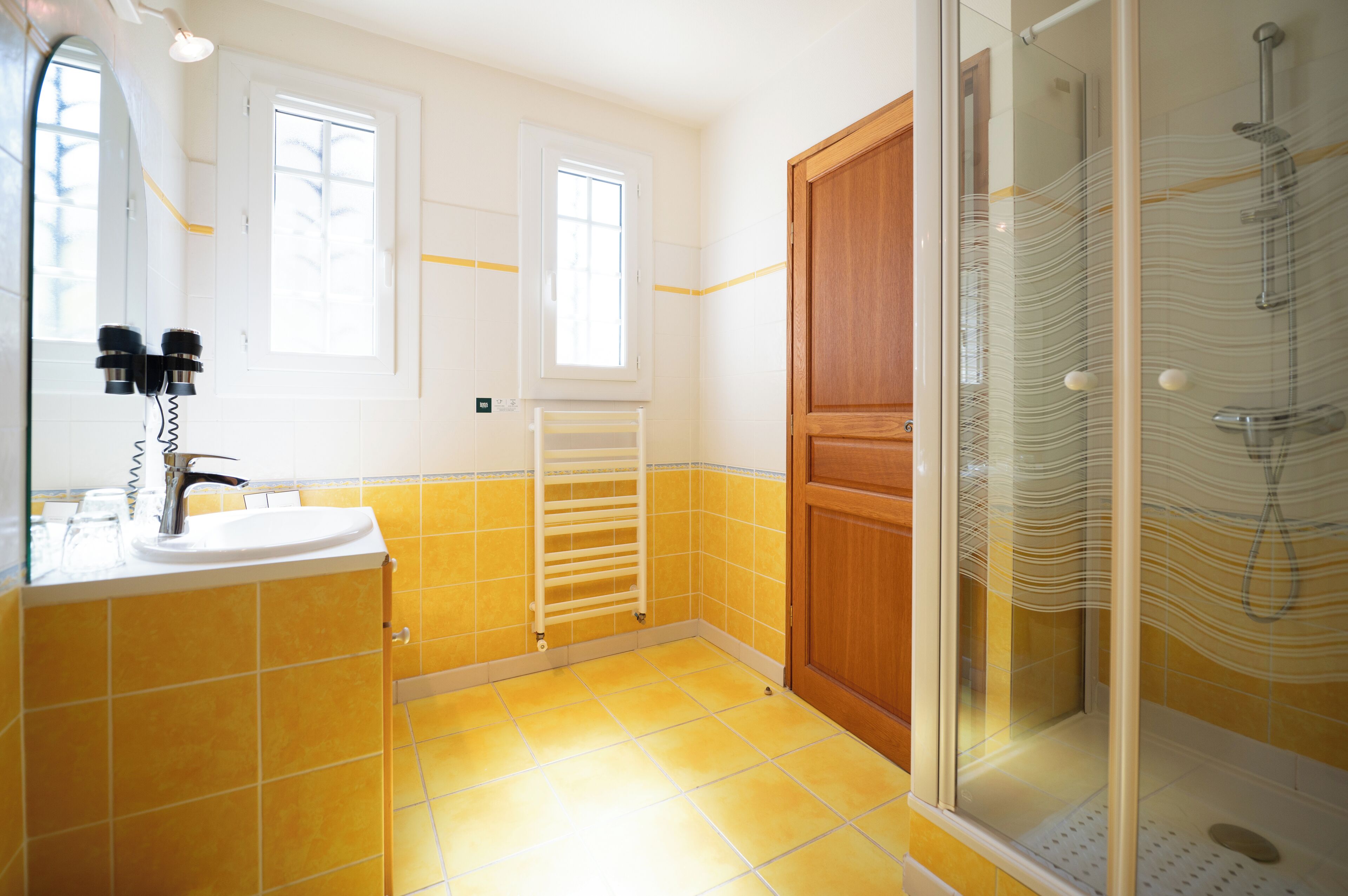 A bathroom with yellow tiles, a sink, a mirror, a window, a door, and a shower enclosure.
