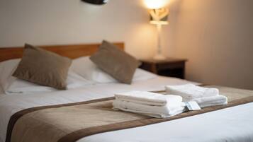 Una cama bien hecha con sábanas blancas y dos almohadas color beige, un cabecero de madera y una mesita de noche con una lámpara.
