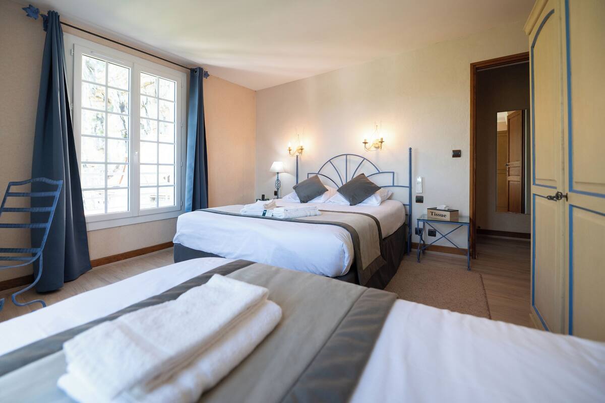 A hotel room with two beds, a window with curtains, a bedside table with a lamp, and a door leading to another room.