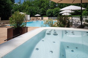 Un jacuzzi con borde de azulejos, rodeado de sillones de descanso y sombrillas, con una piscina y vegetación al fondo.