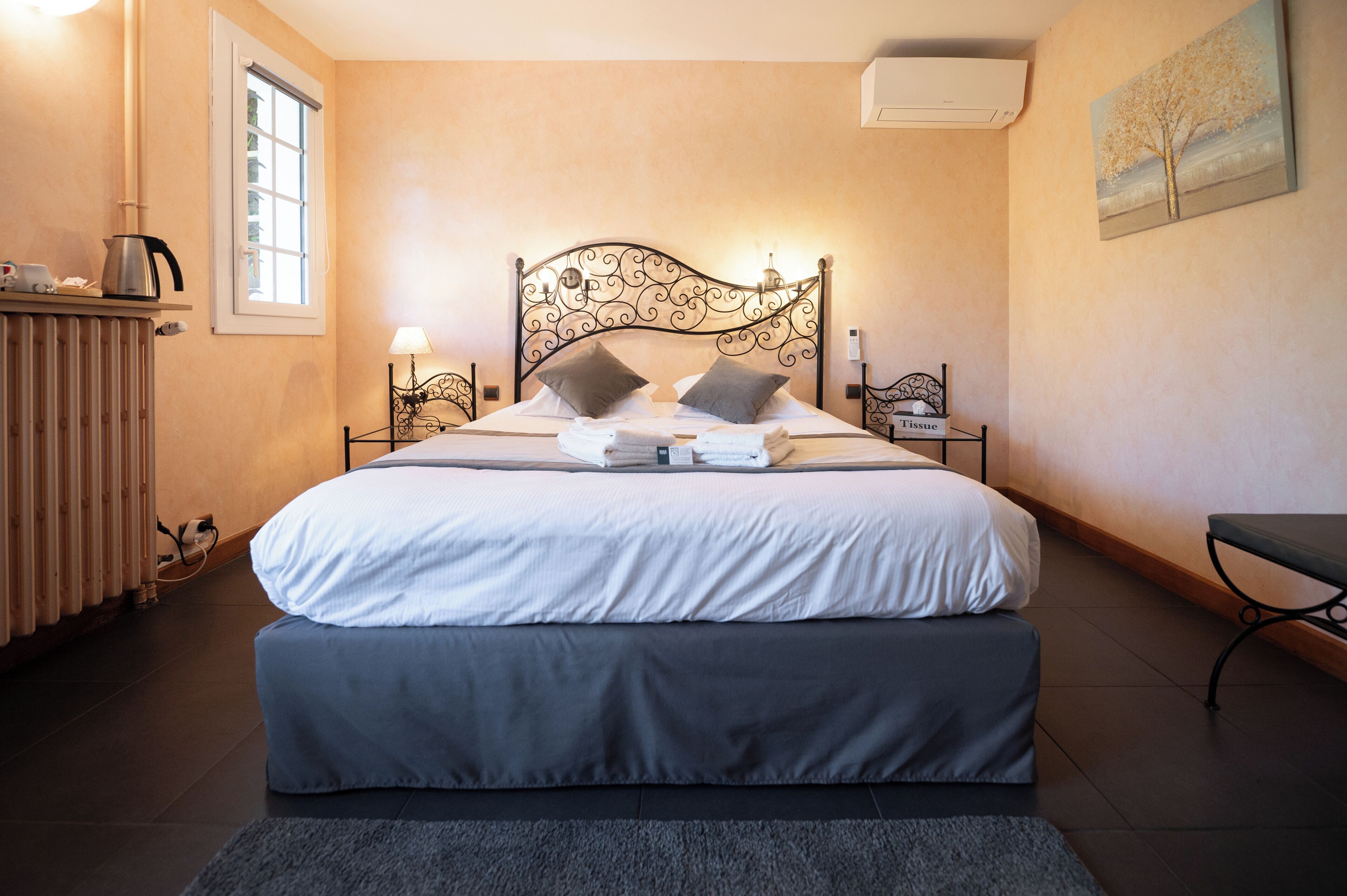 A neatly made bed with a headboard, bedside tables with lamps, a radiator, and a painting on the wall.