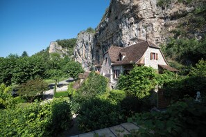 Una casa de piedra con techo pronunciado, rodeada de abundante vegetación y un acantilado.