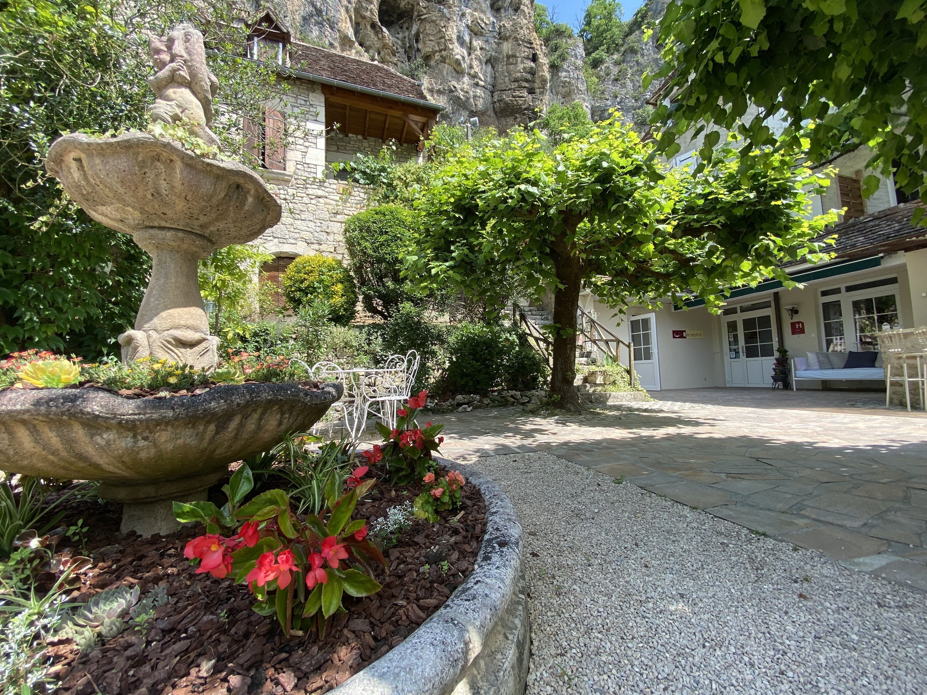 Une fontaine en pierre, une table en métal blanc et un banc en pierre dans un jardin, avec un bâtiment en pierre en arrière-plan.