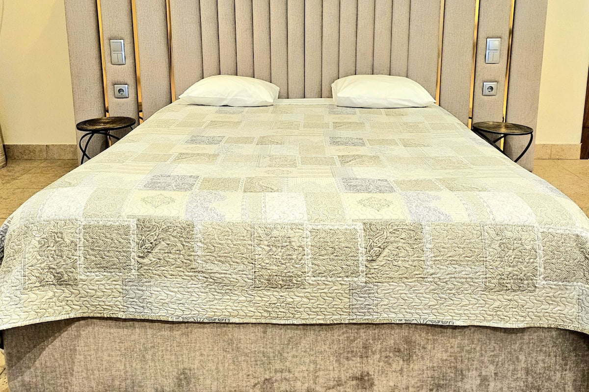 A hotel room with a large bed, two bedside tables, a headboard with vertical slats, and a framed artwork on the wall.