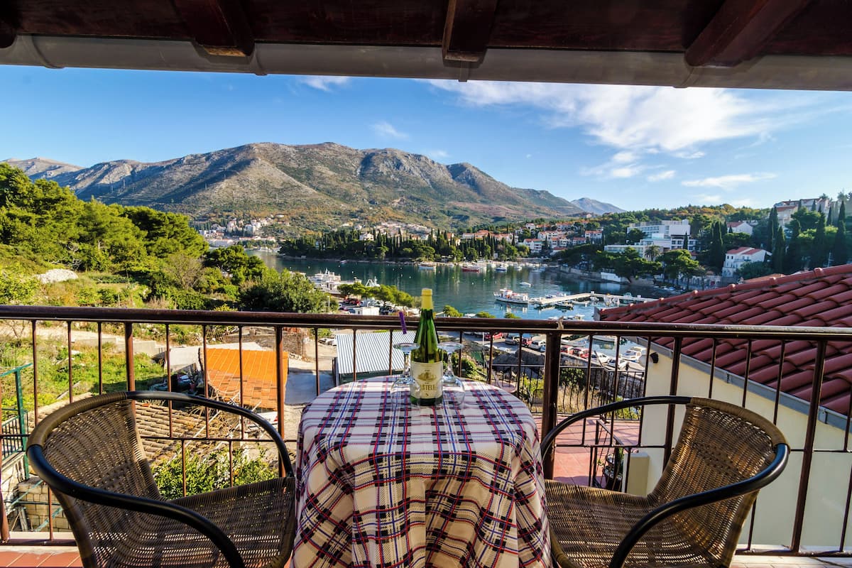 Un balcon avec une table dressée pour deux personnes, donnant sur un lac où naviguent des bateaux, avec une chaîne de montagnes en toile de fond.