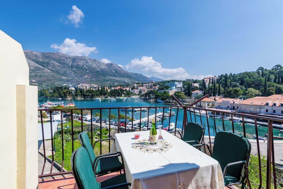 Un balcon avec une table dressée pour le dîner, donnant sur une marina où sont amarrés des bateaux, avec une chaîne de montagnes en arrière-plan.