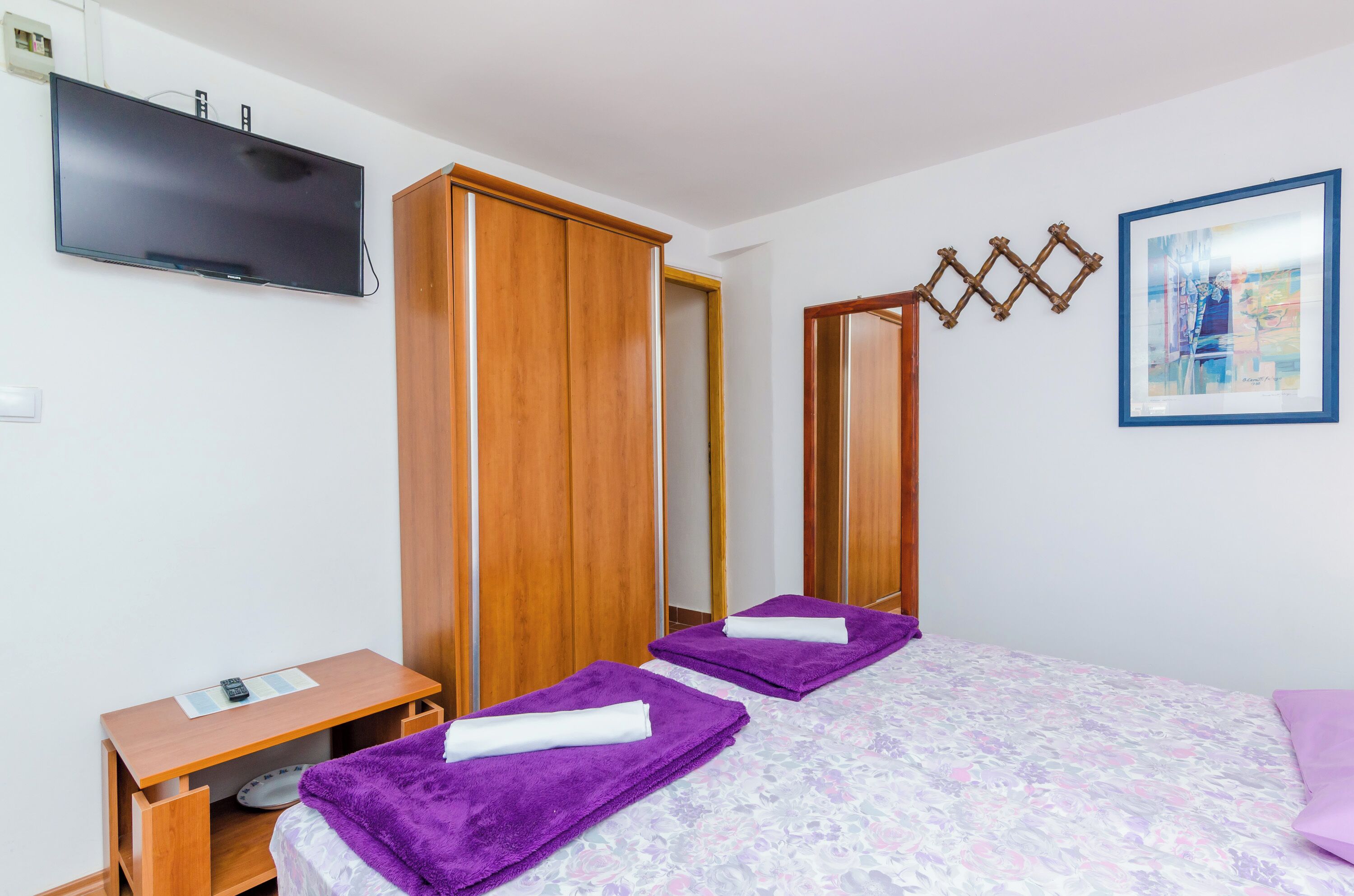 A bedroom with two beds, a wooden wardrobe, a TV mounted on the wall, and a framed artwork.