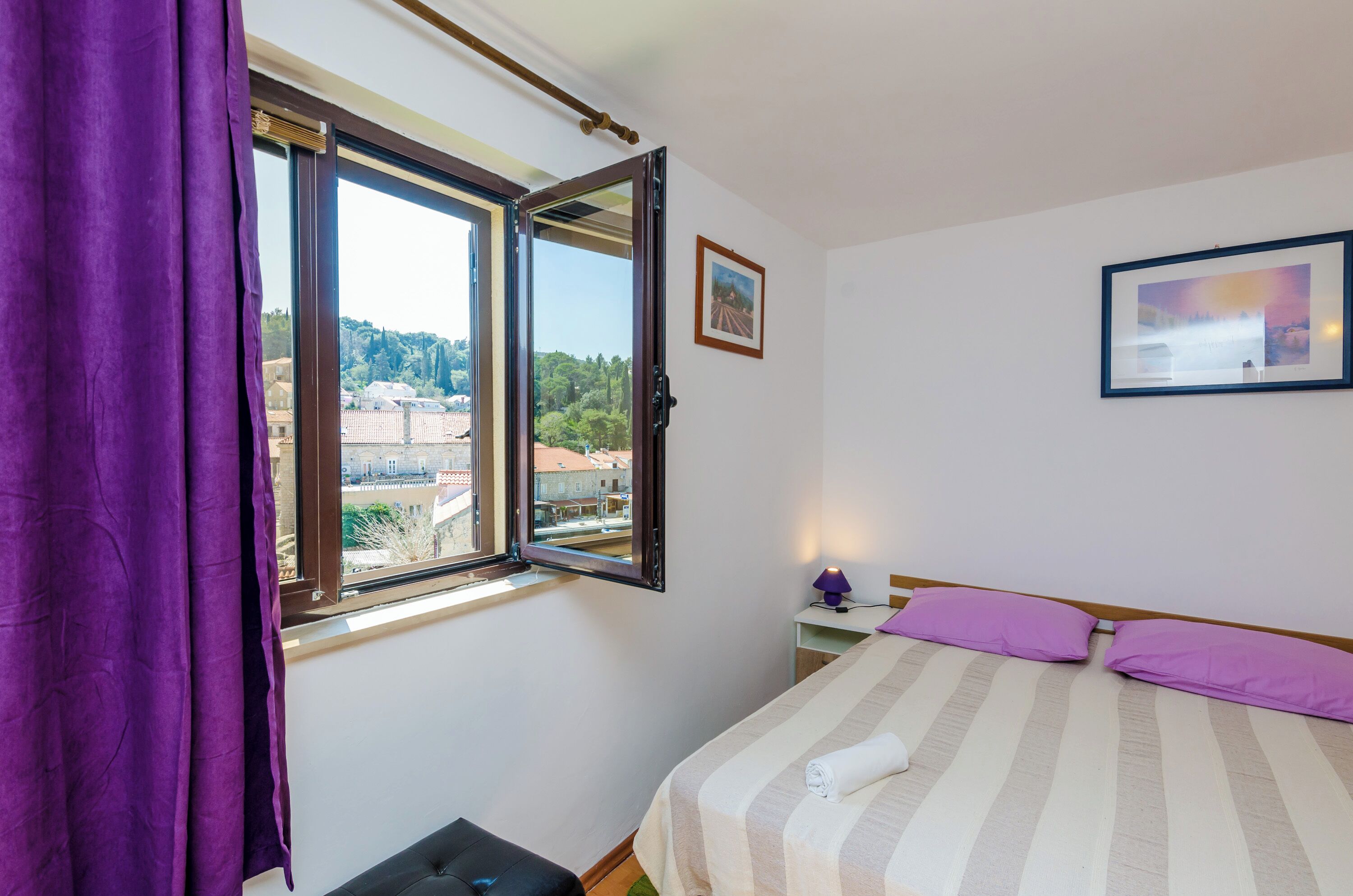 Une chambre à coucher avec un lit, une fenêtre donnant sur des immeubles, un rideau violet et un tableau encadré accroché au mur.