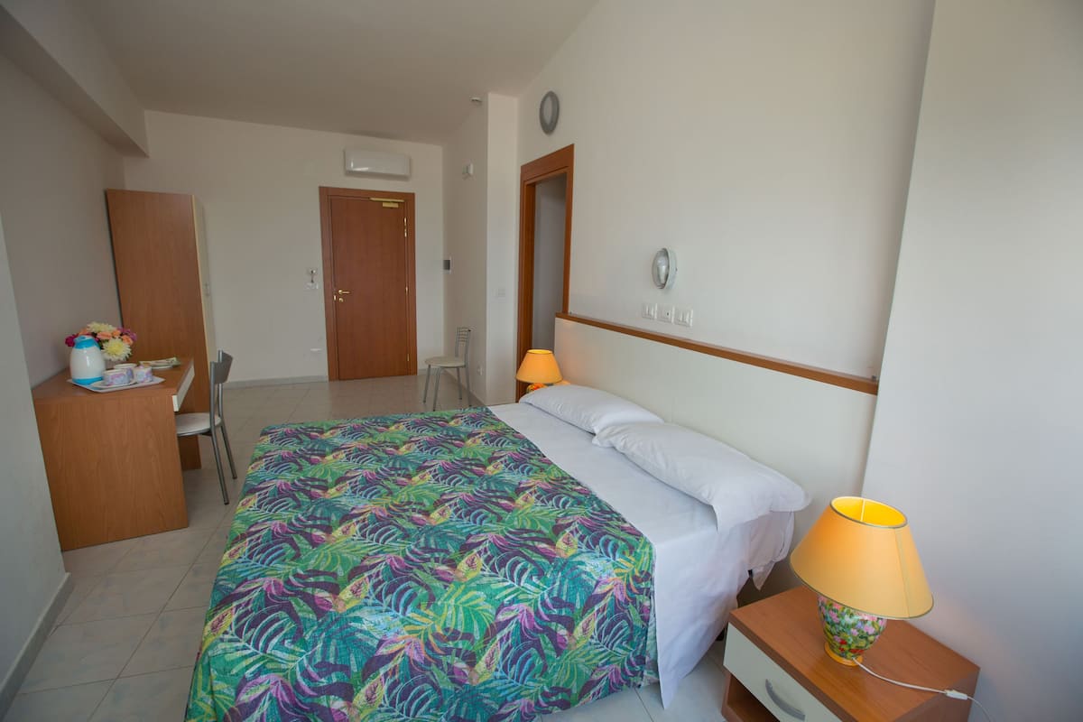 A hotel room with a bed, bedside table with a lamp, a desk with a chair, and a wardrobe.
