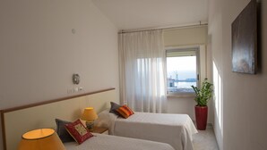 Una camera d'albergo con due letti, un comodino, una lampada, una pianta in vaso e una finestra con vista.