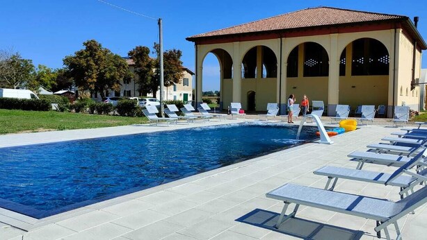 A swimming pool with lounge chairs and a building with arches in the background. - Corte Galvana (Cento)