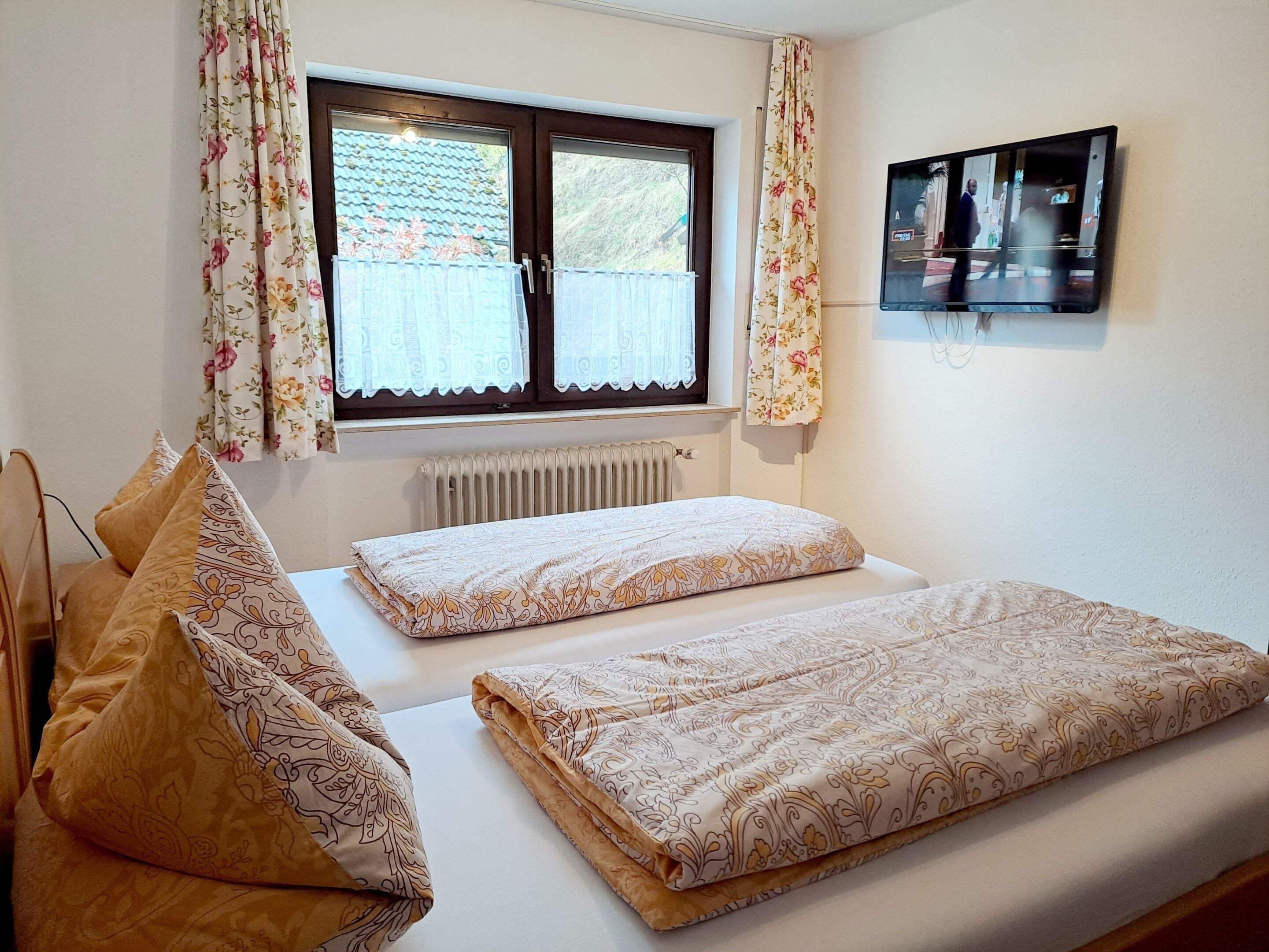 A bedroom with two beds, floral curtains, a radiator, and a TV on the wall.