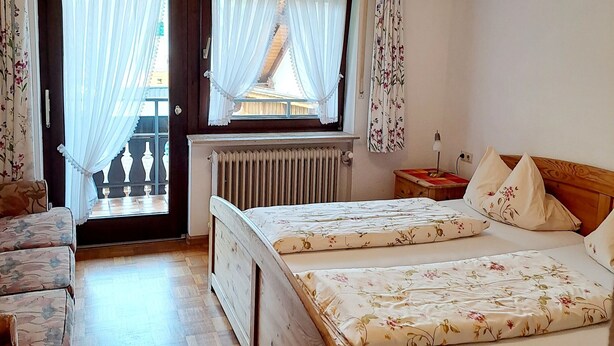 A bedroom with two wooden beds, a wooden floor, a window with curtains, a radiator, and a ceiling light fixture. - Pension Schlossbergblick (Simonswald)