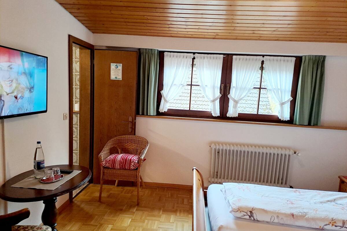 A room with a wooden floor, a bed with floral bedding, a wicker chair, a table with a bottle and glasses, a framed certificate on the wall, and a window with curtains.