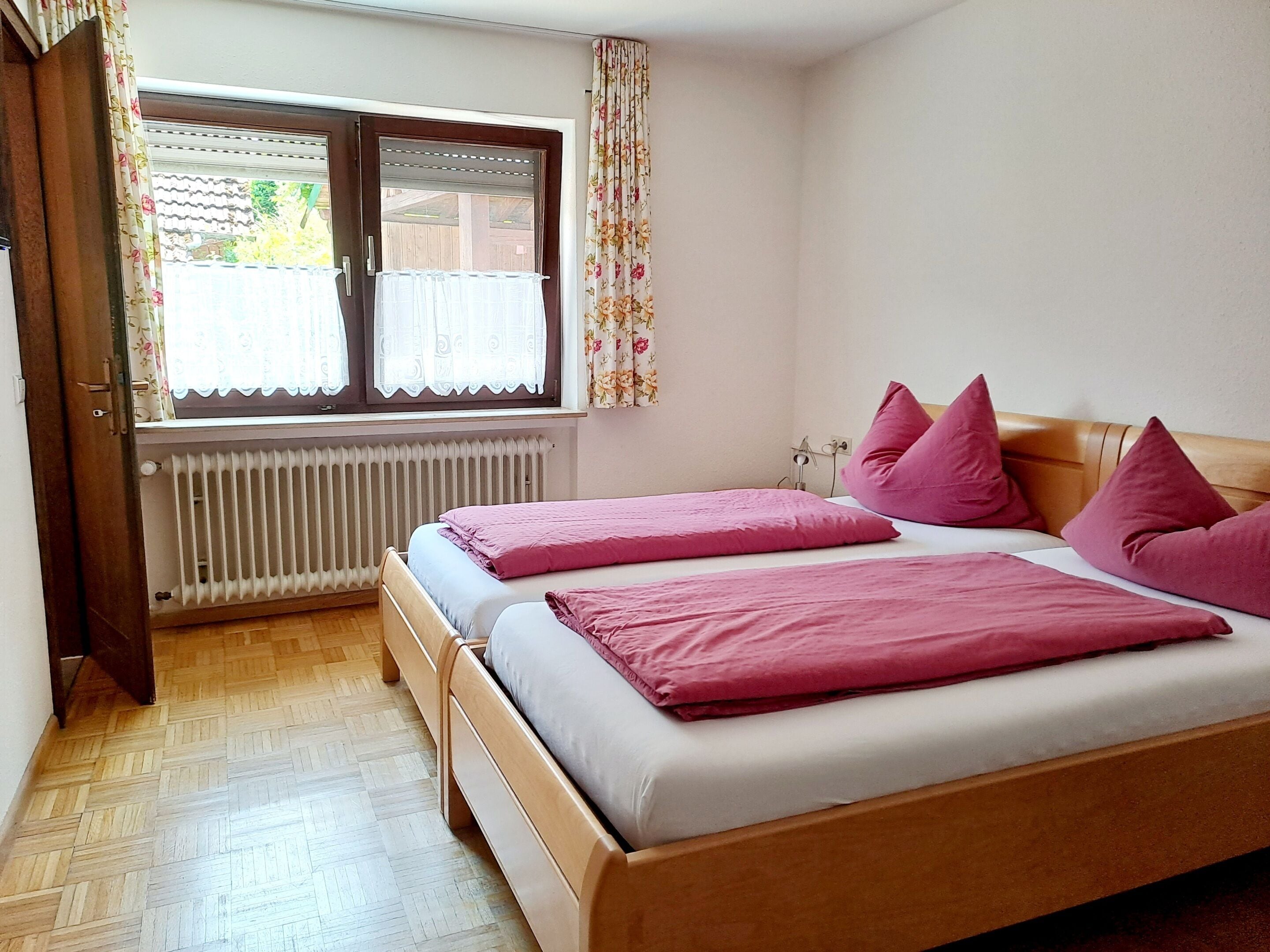 Een slaapkamer met twee bedden, een radiator, een raam met bloemen gordijnen, en een houten vloer.