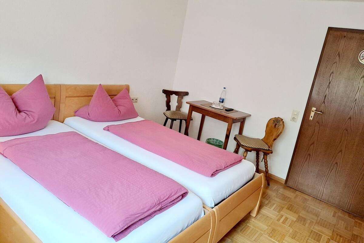 A double bed with pink bedding, a wooden headboard, a small wooden table with a water bottle, and a chair.