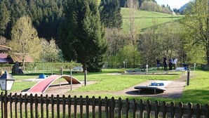 A park with playground equipment, a wooden fence, and a backdrop of trees and hills. - Pension Schlossbergblick (Simonswald)