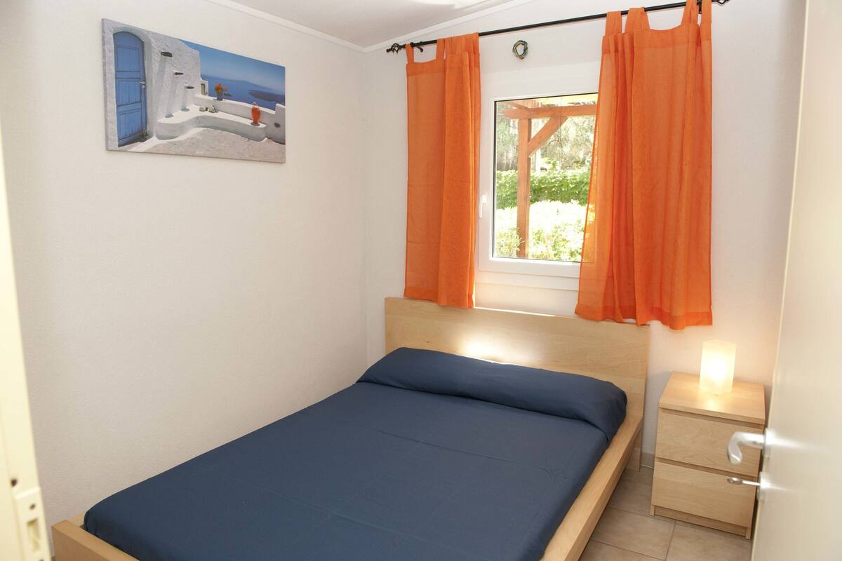 Una cama individual con cabecera de madera, una mesita de noche con lámpara y una ventana con cortinas anaranjadas.