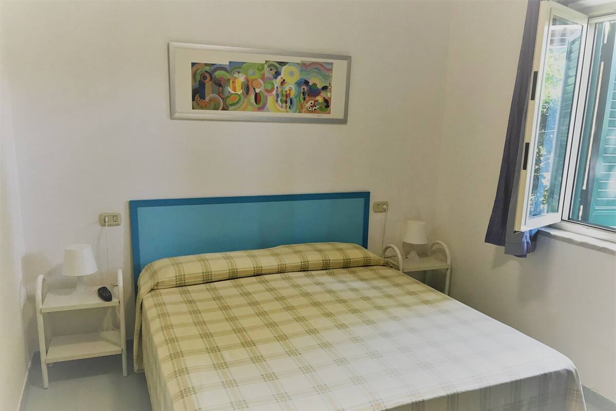 Un dormitorio con cama, dos mesitas de noche, una mesita auxiliar, una ventana con cortinas y un cuadro colgado en la pared.