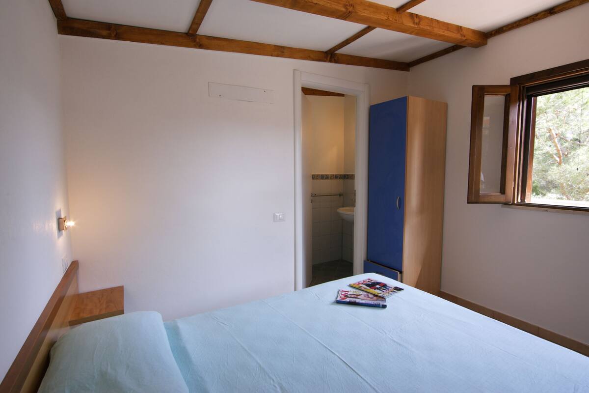 Un dormitorio con cama, ventana, puerta que da a un baño y una viga de techo de madera.