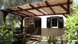 A covered patio area with a table and chairs, a small garden with flowers, and a building with a window and shuttered door. - Villaggio Residence Torre Saracena - Campsite (Camerota)