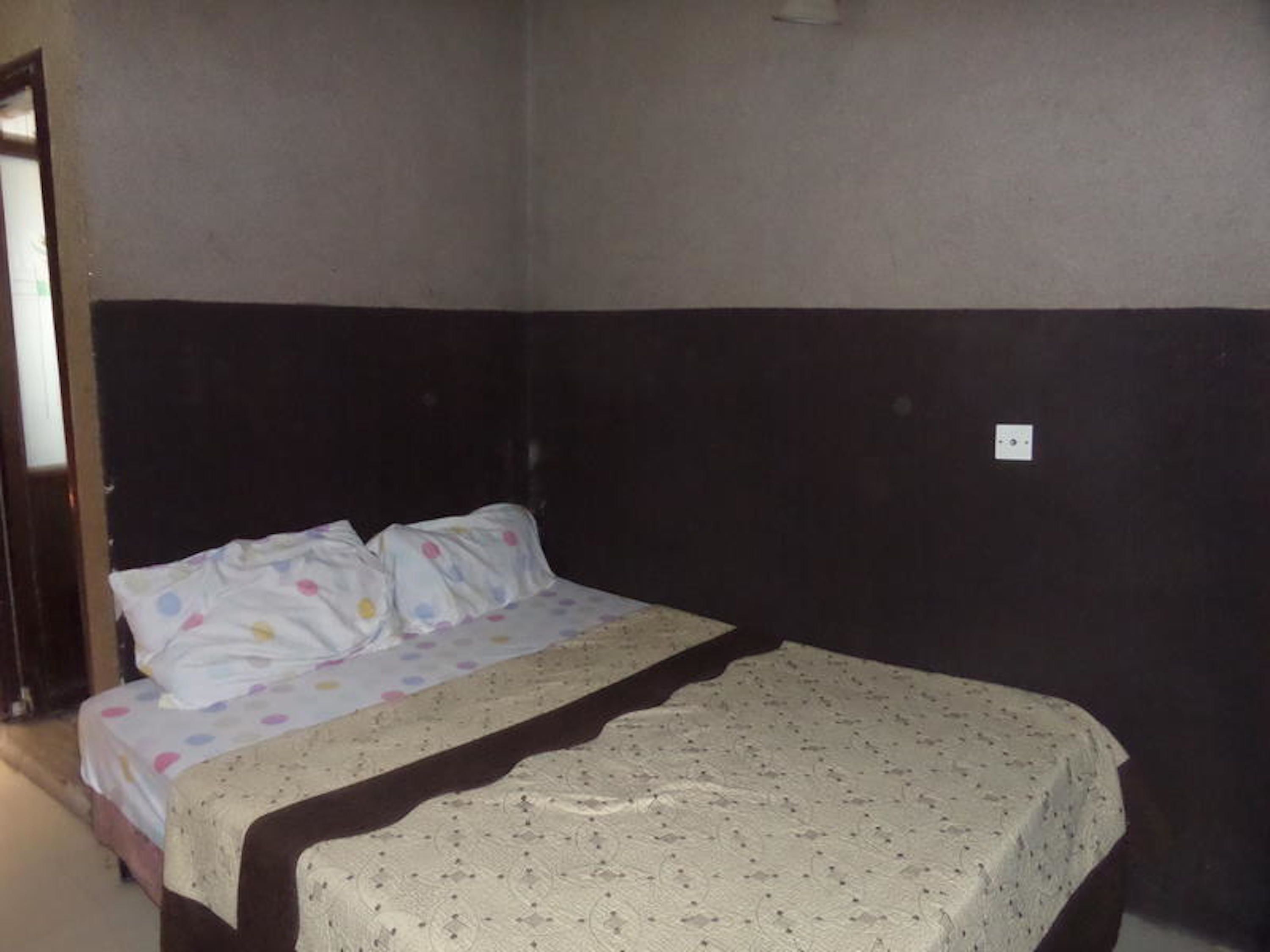 A single bed with a patterned bedspread and pillows, a dark headboard, and a plain wall with a single electrical outlet.