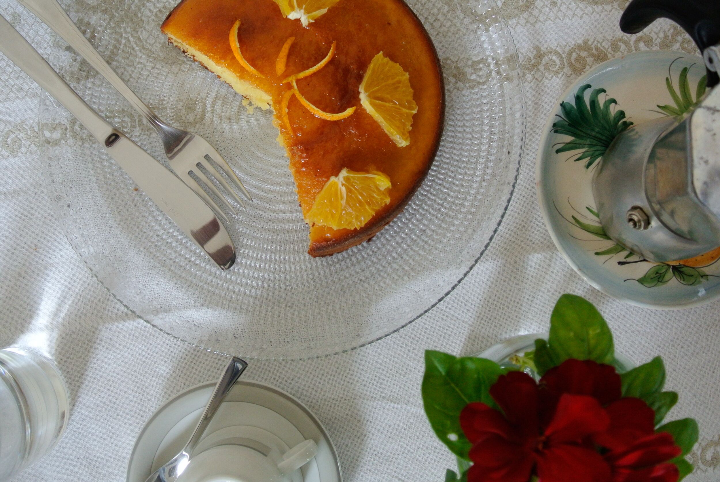 A slice of cake on a plate with a fork beside it, a teapot, and a flower on a tablecloth.