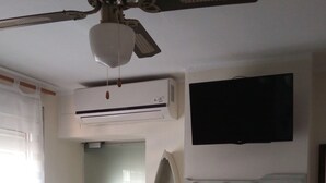 A room with a ceiling fan, air conditioning unit, a flat-screen TV mounted on the wall, a small table with two chairs, and a door leading to another room. - Pensión el Hidalgo (Calpe)