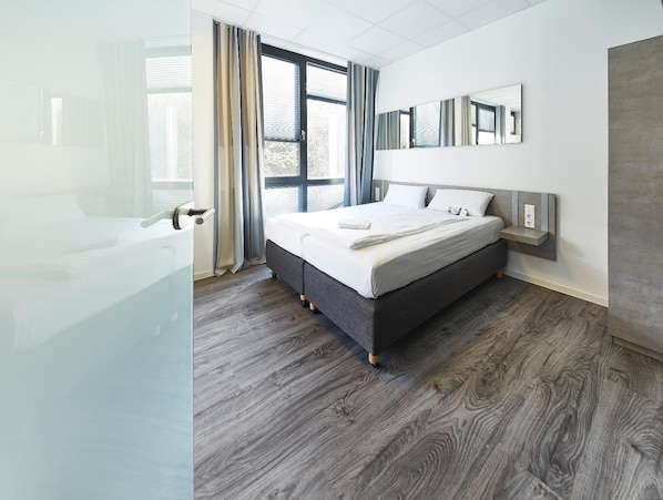 A modern bedroom with a large bed, a bedside table, a mirror, and a view of the outdoors through large windows. - Road House Hotel (Paderborn)