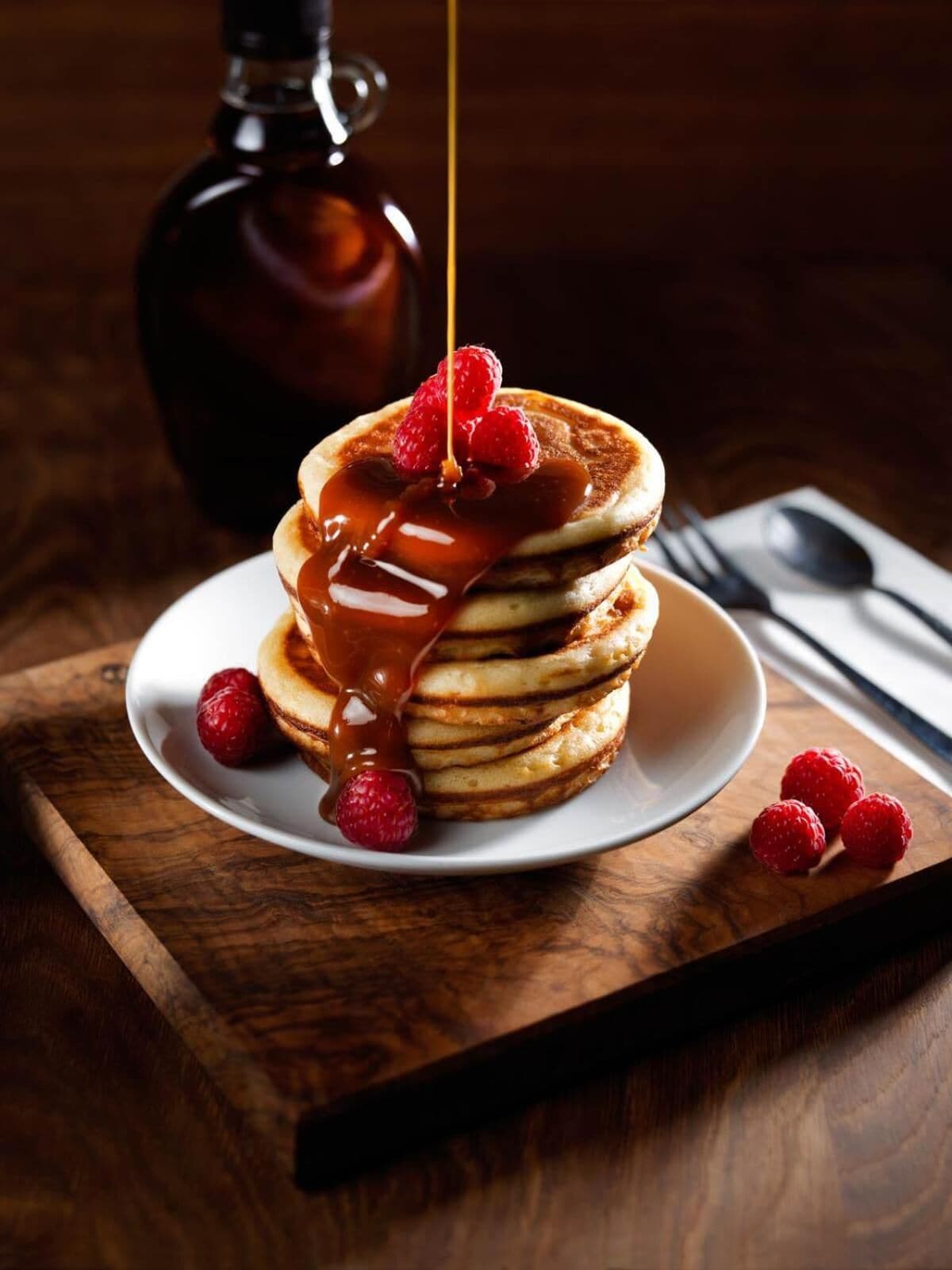 Una pila di pancake con sciroppo e lamponi su un piatto bianco, posizionata su un tavolo di legno con una bottiglia e utensili sullo sfondo.