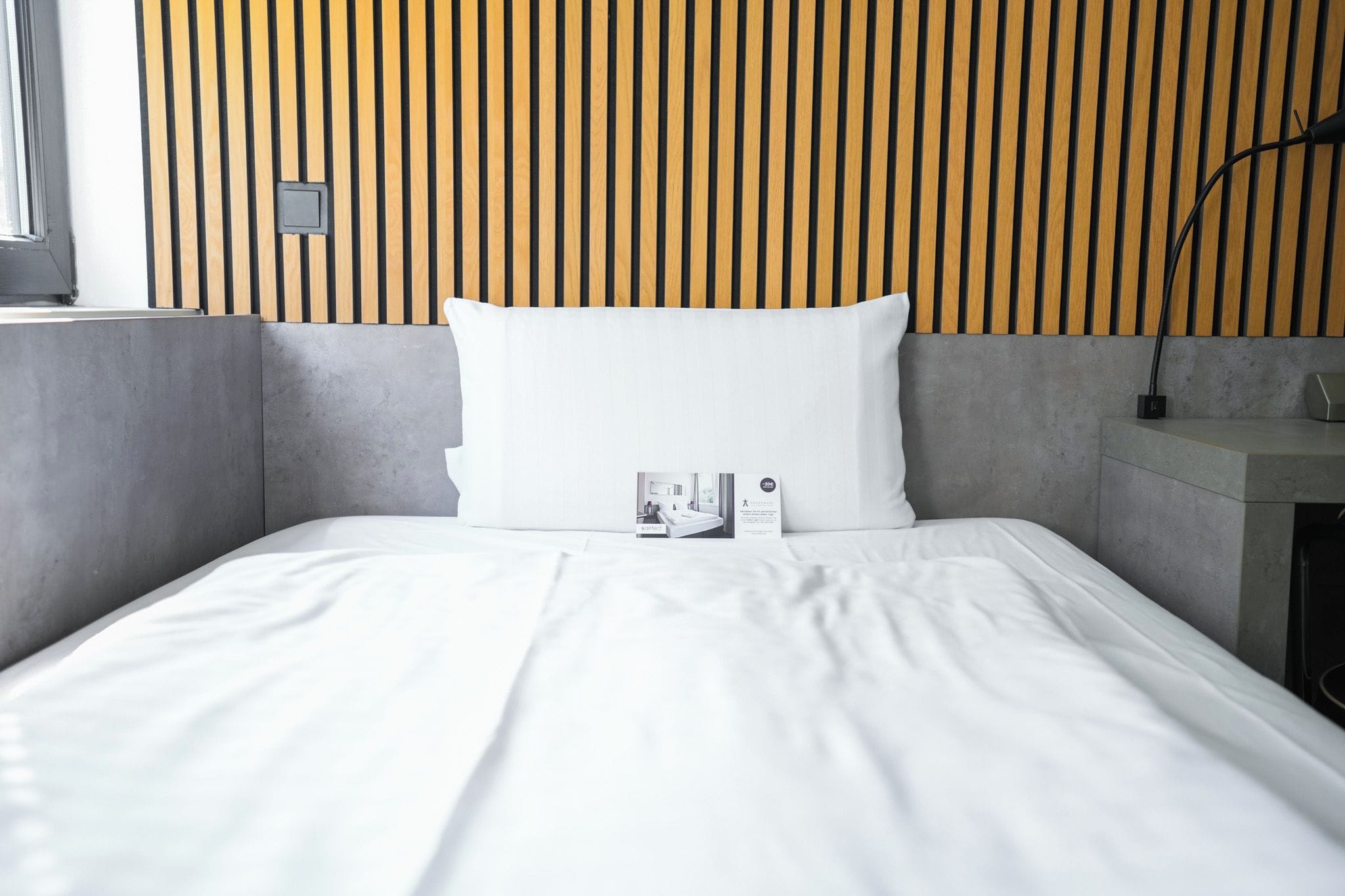 A neatly made bed with white linens, a pillow, and a bedside table with a lamp and a small electronic device.
