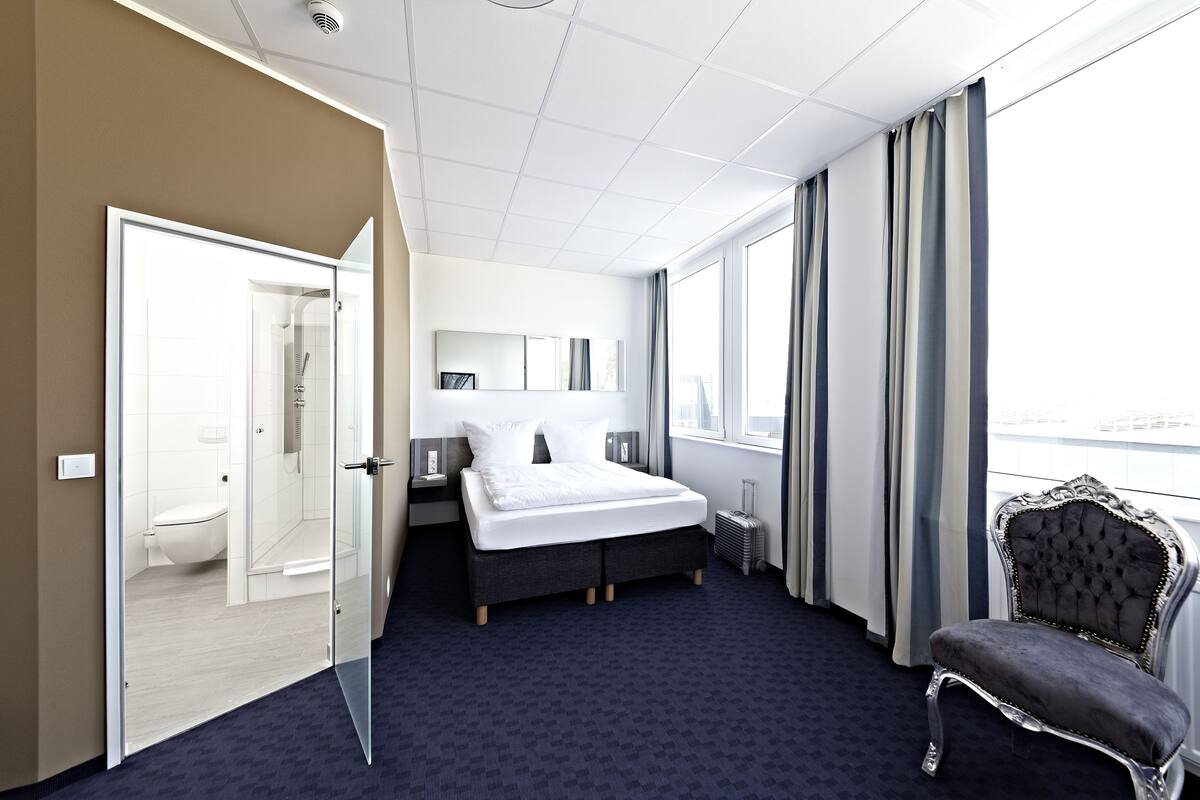 Una habitación de hotel moderna con una cama grande, un área de descanso con una silla, ducha con cerramiento de vidrio y vista a la ciudad.