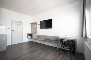 A modern, minimalistic room with a bench, a desk, a chair, a flat-screen TV mounted on the wall, and a door with a lock and a calendar. - Road House Hotel (Paderborn)