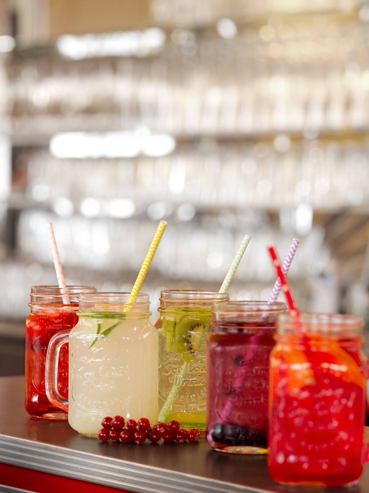 A selection of colorful beverages in mason jars with straws, some garnished with fruits, displayed on a shelf.