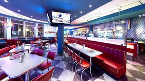 Un restaurant à la décoration rétro, avec des banquettes et des tables, un grand écran de télévision au centre et un mur orné de motifs.