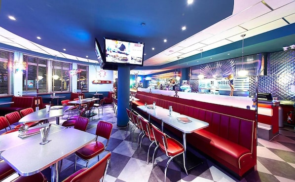 A retro-themed restaurant with booths and tables, a central TV screen, and a patterned wall.