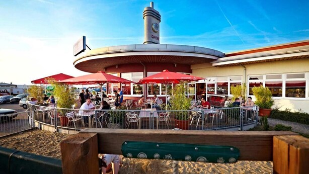 Un espace extérieur aménagé pour les repas, avec des tables et des parasols rouges, entouré d’une clôture, et une tour de l’horloge en arrière-plan.
