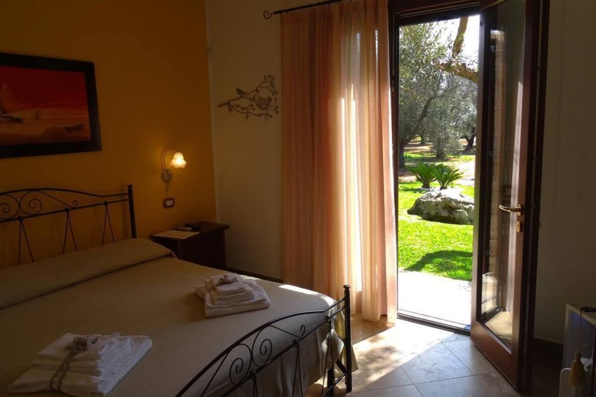 Ein Schlafzimmer mit Bett, Nachttisch, Lampe und Blick auf einen Garten durch eine offene Tür.