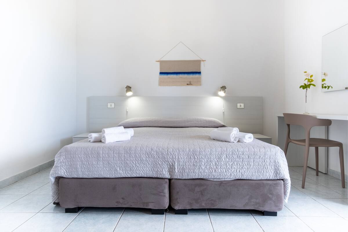 A modern bedroom with a large bed, bedside lamps, a desk, and a hanging artwork.