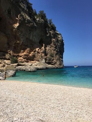 A beach with clear turquoise water, rocky cliffs, and a boat in the distance.
