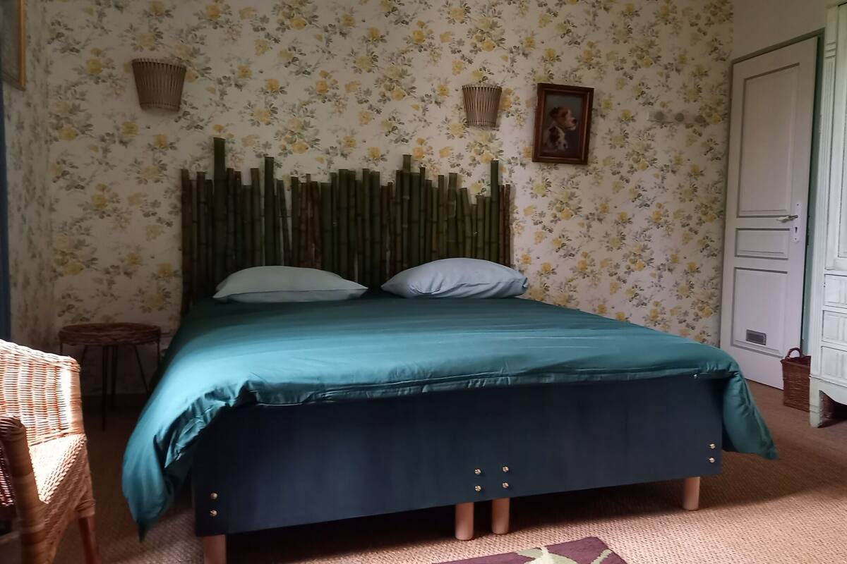 Sänky, jossa on vihreä päiväpeite, kaksi tyynyä, puinen pääty, rottinkituoli, pieni pyöreä pöytä ja valkoinen lipasto.