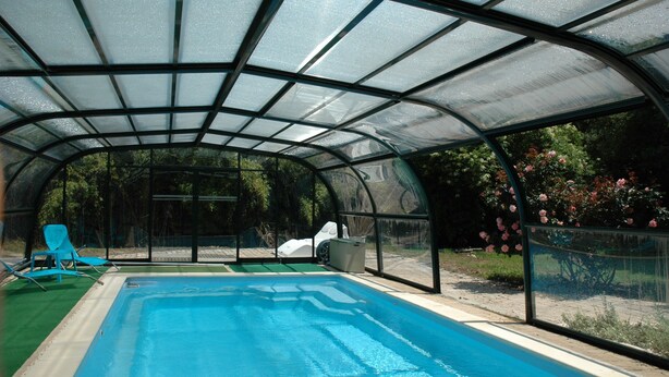 A covered swimming pool with a glass roof, surrounded by greenery and a white car. - La Palombiere (Lucmau)